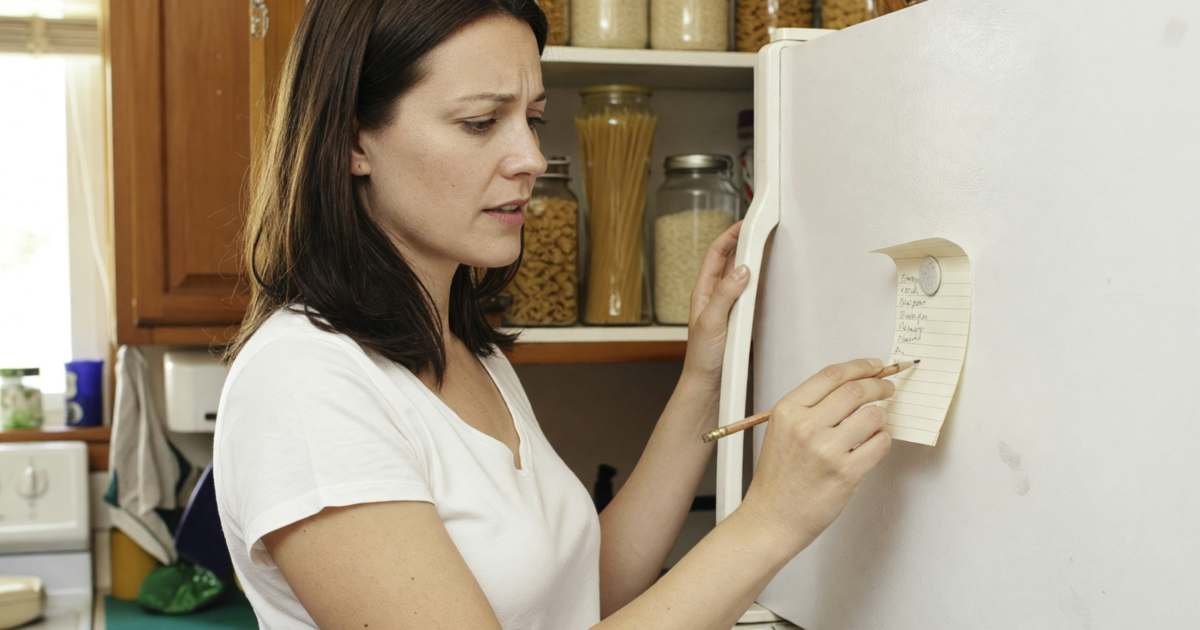 A woman writing a grocery list on a small magnetic notepad stuck to her fridge while checking the pantry shelves, illustrating a reusable Canadian grocery list tied to flyers and loyalty offers.