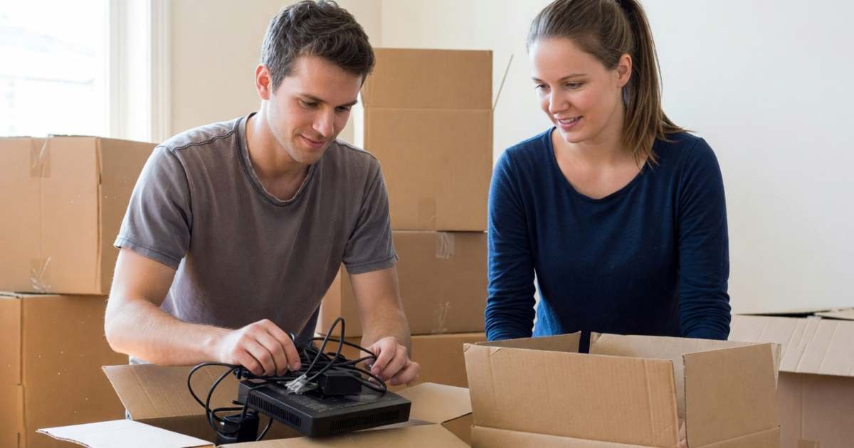 A young couple packing up a modem and cables into a moving box among other packed boxes, illustrating cancelling an internet contract early in Canada.