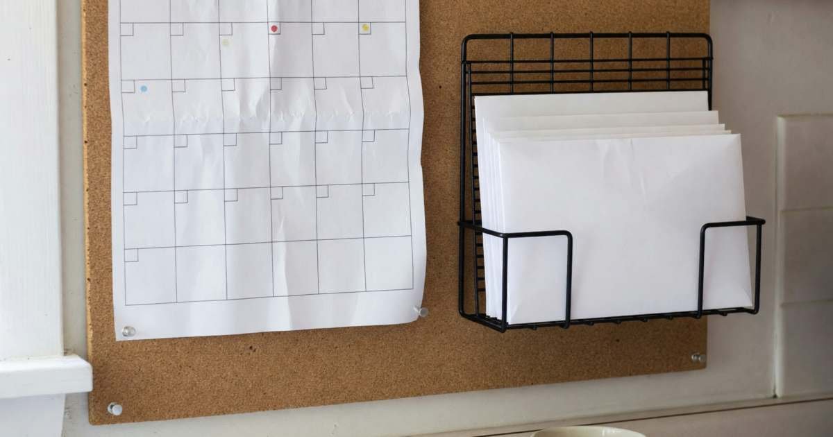A paper wall calendar with a blank grid pinned on a kitchen bulletin board, some squares marked with coloured dots, beside a few envelopes in a wire organiser, illustrating building a cashflow calendar for paydays and bills in Canada.