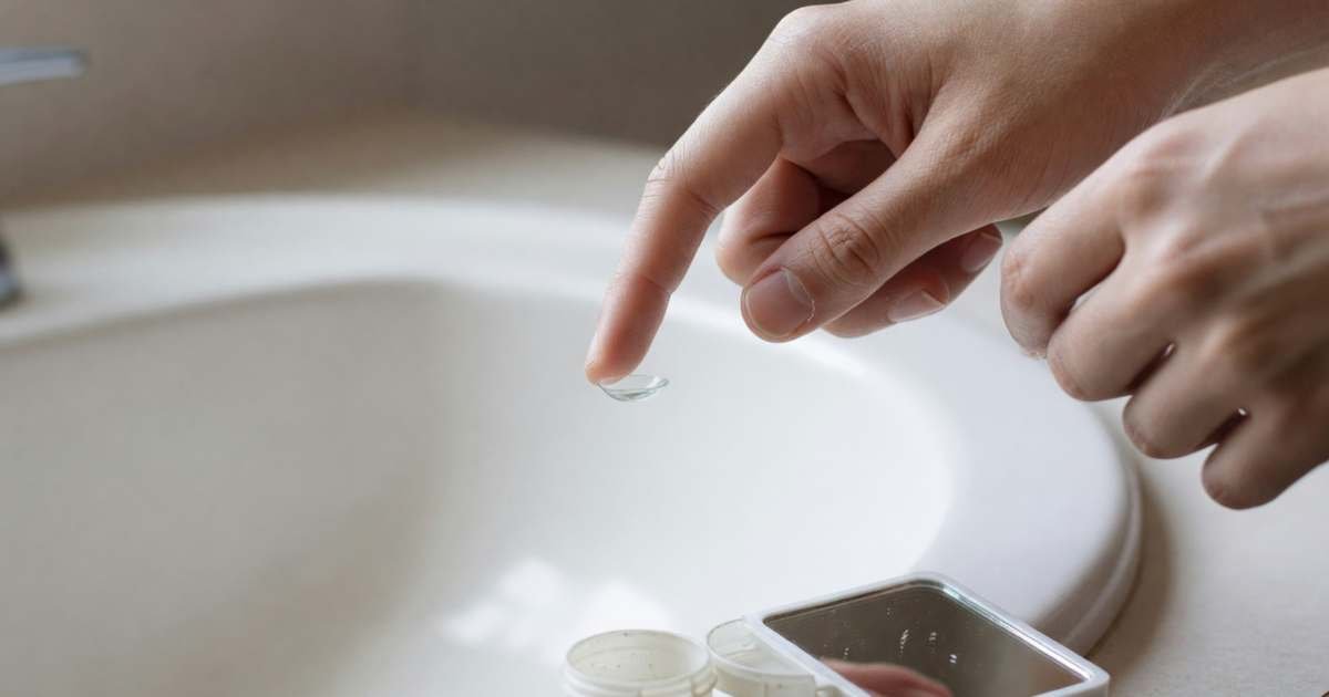 Fingers holding a contact lens on a fingertip above a bathroom sink, illustrating annual cost of contact lenses in Canada.