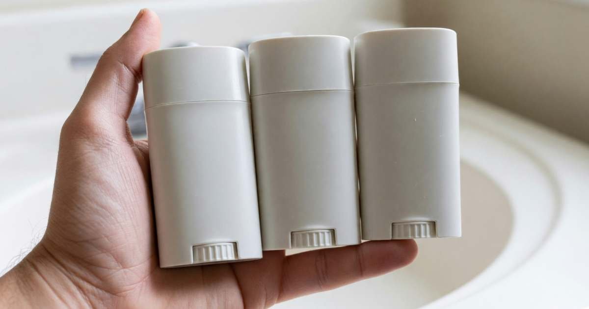 A hand holding three plain deodorant sticks side by side over a bathroom counter, illustrating deodorant costs, natural versus regular versus store brand.