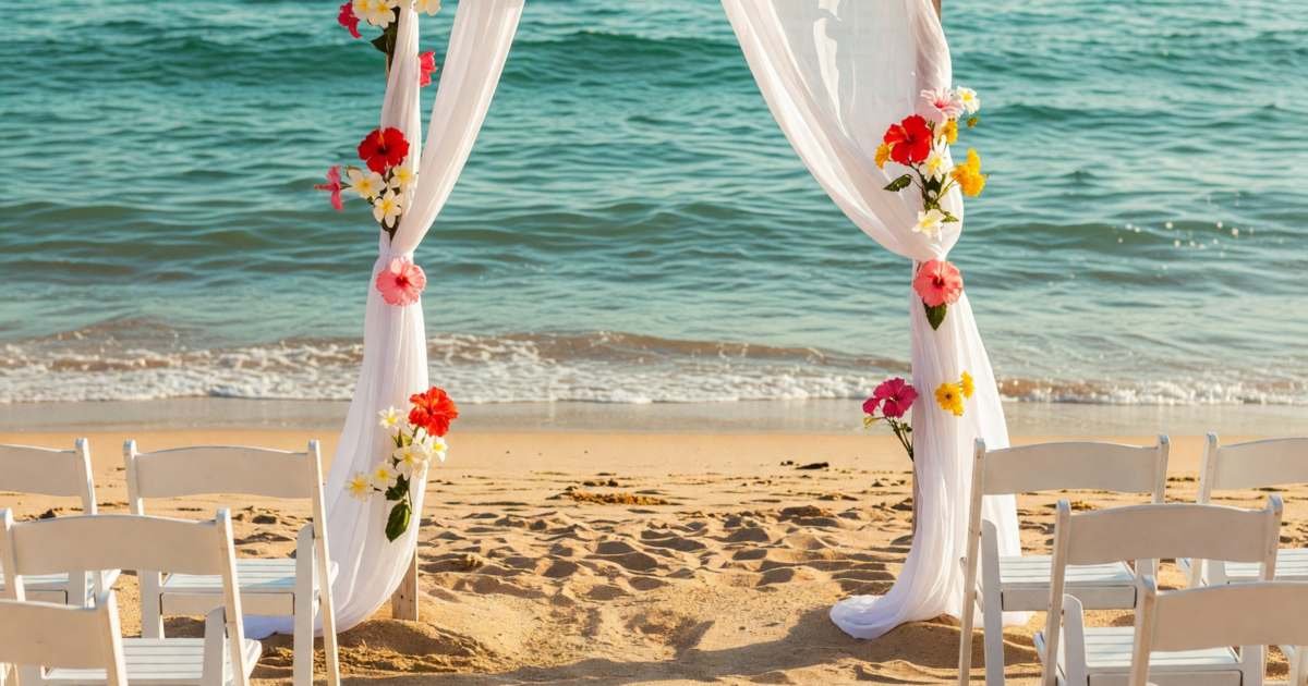 A small beach wedding arch draped with white fabric and flowers facing the ocean, rows of white chairs, illustrating destination wedding costs for couples and guests.