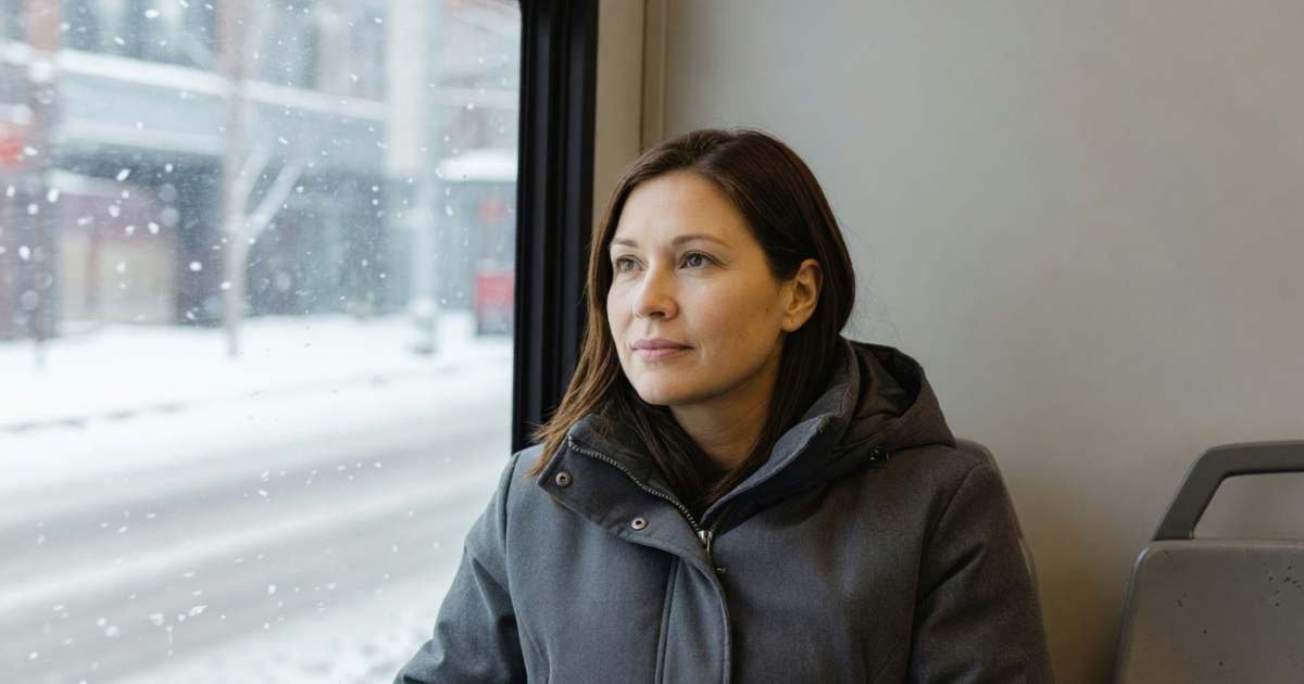 A woman in a winter coat sitting by the window on a city bus, looking out with a calm expression, illustrating Toronto's Fair Pass transit discount and fare capping.