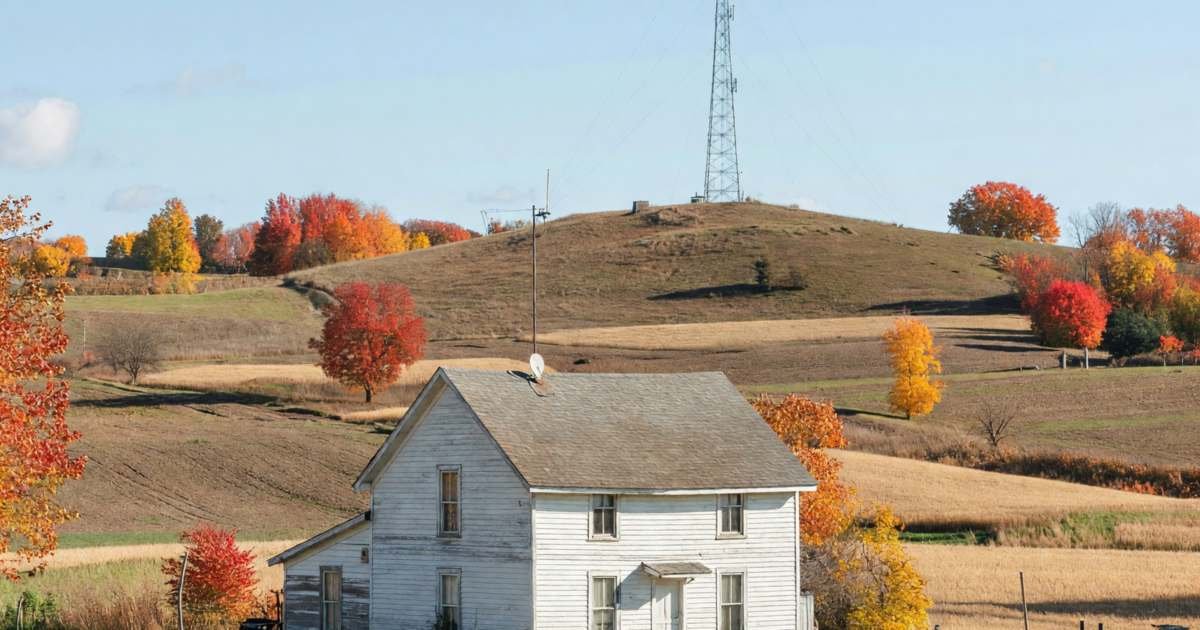 A farmhouse with a small fixed wireless antenna on the roof and a radio tower on a distant hill, illustrating fixed wireless internet providers in rural Canada.