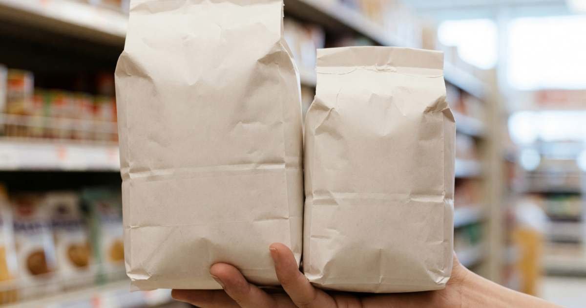 Close-up of a hand holding two different-sized plain bags of rice side by side in a grocery aisle to compare them, illustrating reading unit prices at Canadian grocery stores.