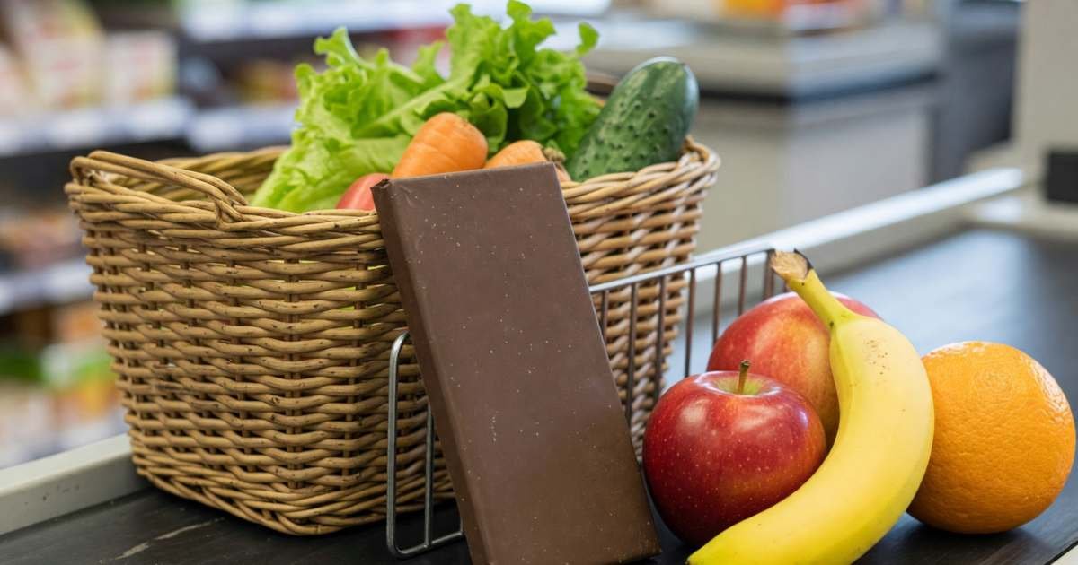 A chocolate bar sitting back on a rack beside fruit and a basket of vegetables at a grocery checkout, illustrating stopping impulse grocery spending in Canada.