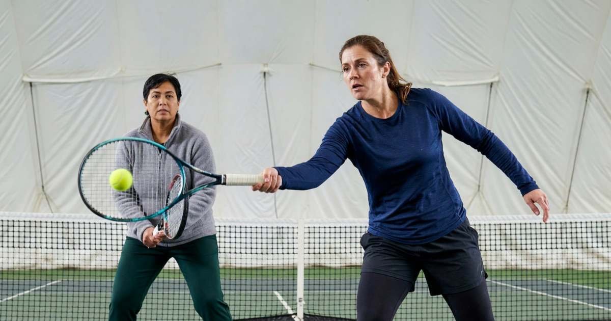 Two women playing doubles-style rallies on an indoor tennis court under a white air-supported dome, illustrating indoor tennis costs in Canada's winter.