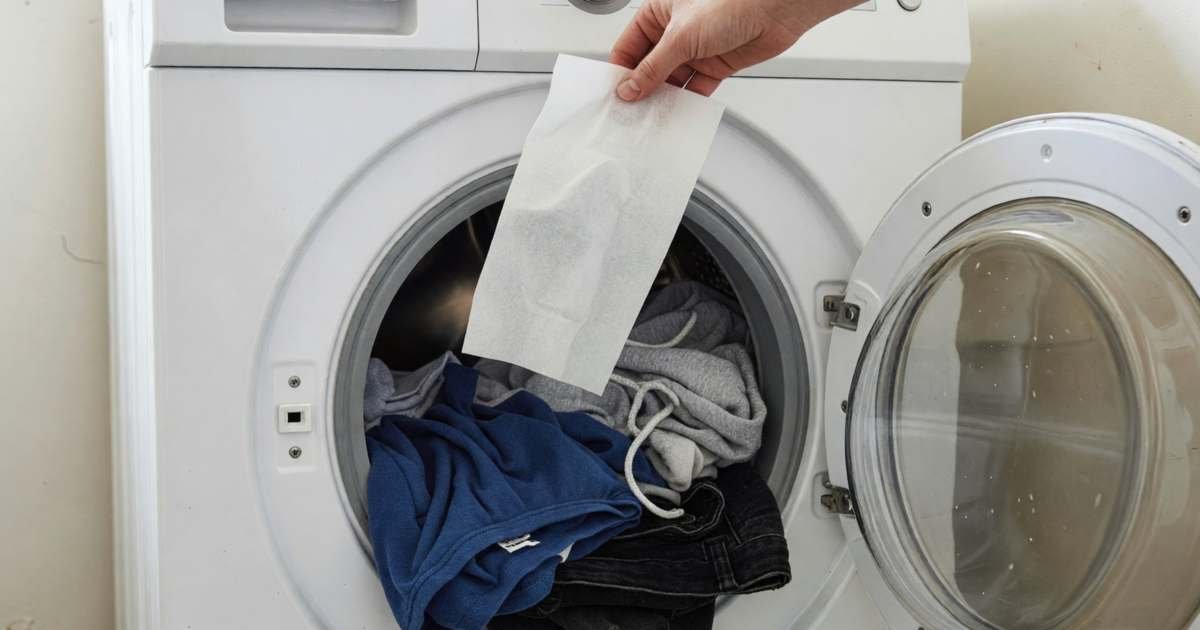 A hand dropping a thin detergent sheet into a washing machine drum full of clothes, illustrating laundry detergent sheets cost per load.