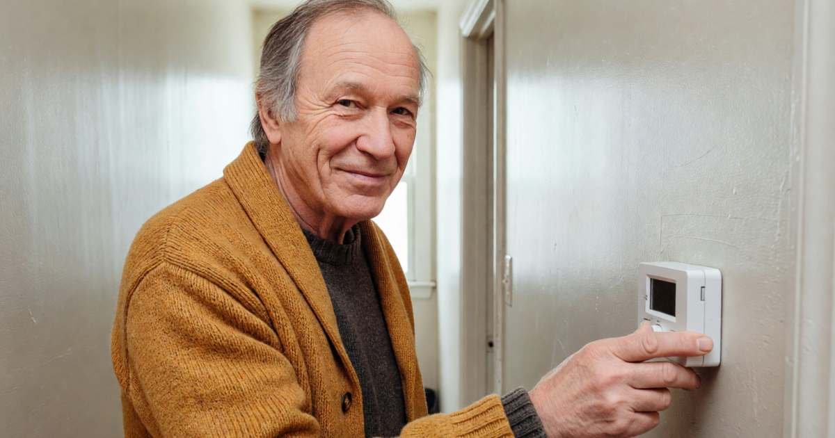 An older man in a warm cardigan turning down a plain wall thermostat in his hallway, illustrating lowering the winter heating bill across Canadian provinces.