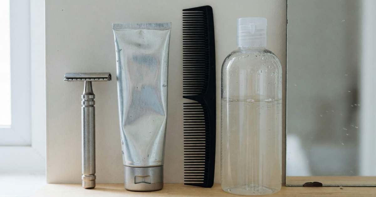 A short row of men's grooming basics on a shelf, a razor, shaving cream, a comb and a bottle of lotion, illustrating men's grooming on a budget.