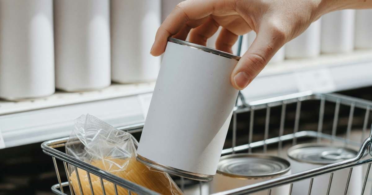 Close-up of a hand placing a plain unlabelled can of tomatoes into a shopping basket already holding pasta and beans, illustrating switching to Canadian store brands like No Name, PC and Compliments.