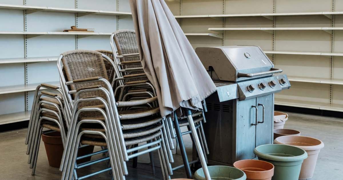 An end-of-season clearance display in a garden centre with stacked patio chairs, a folded patio umbrella, a barbecue and a few garden planters grouped together, half the shelves behind already empty, illustrating a seasonal clearance calendar for the best month to buy in Canada.