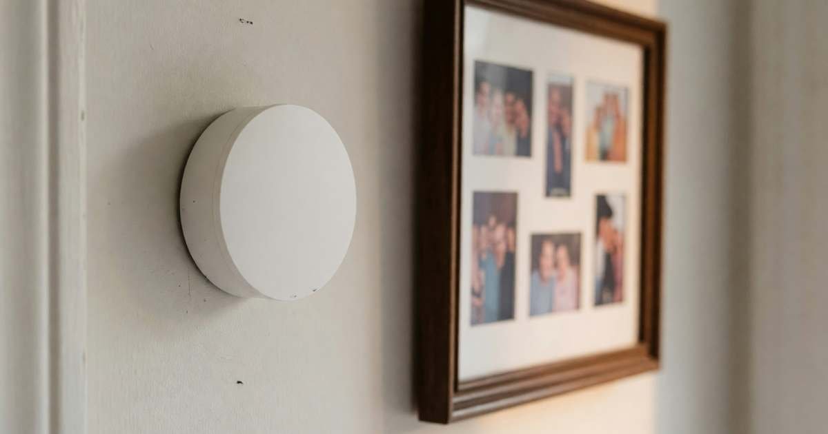 A plain round smart thermostat on a hallway wall beside a family photo frame with blurred photos, illustrating smart thermostat rebates from Canadian utilities.