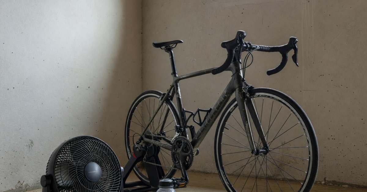A road bike mounted on a smart trainer in a basement corner with a fan, illustrating smart trainer versus spin classes.