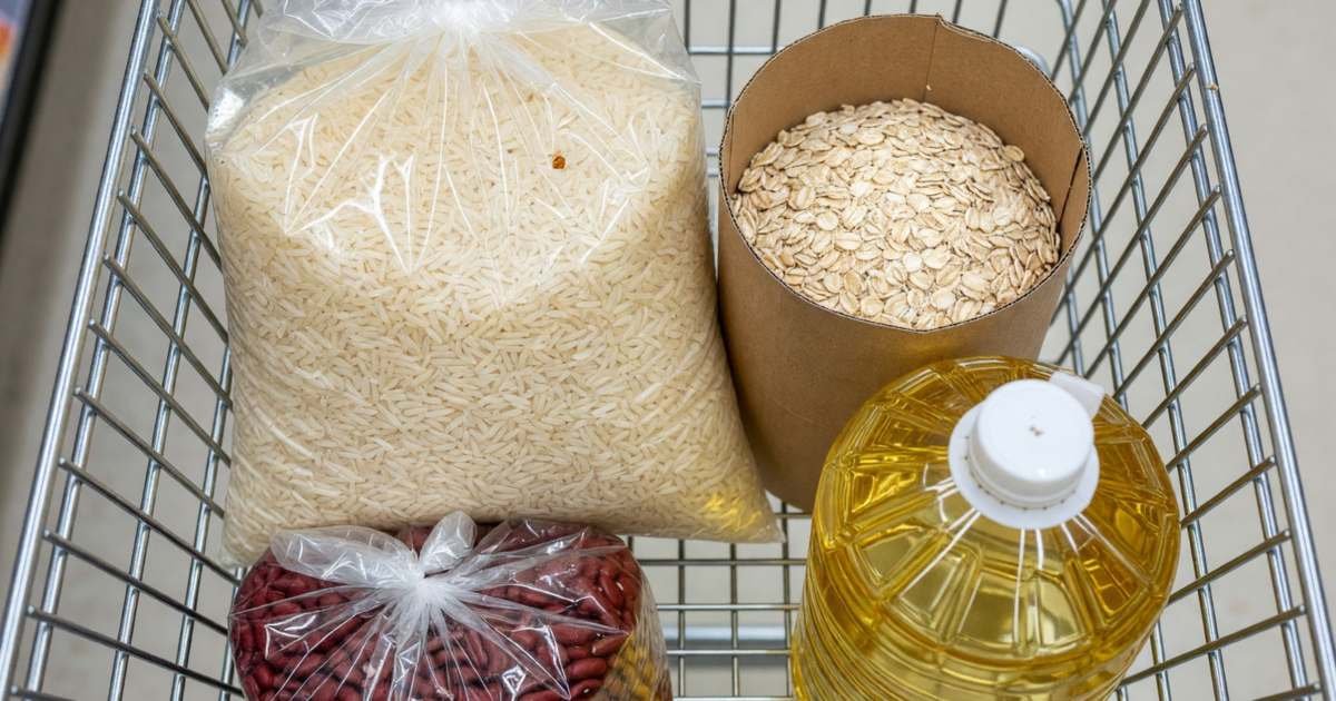 A grocery cart holding a bag of rice, dried beans, oats and a jug of cooking oil, illustrating keeping a staple price book for Canadian groceries.