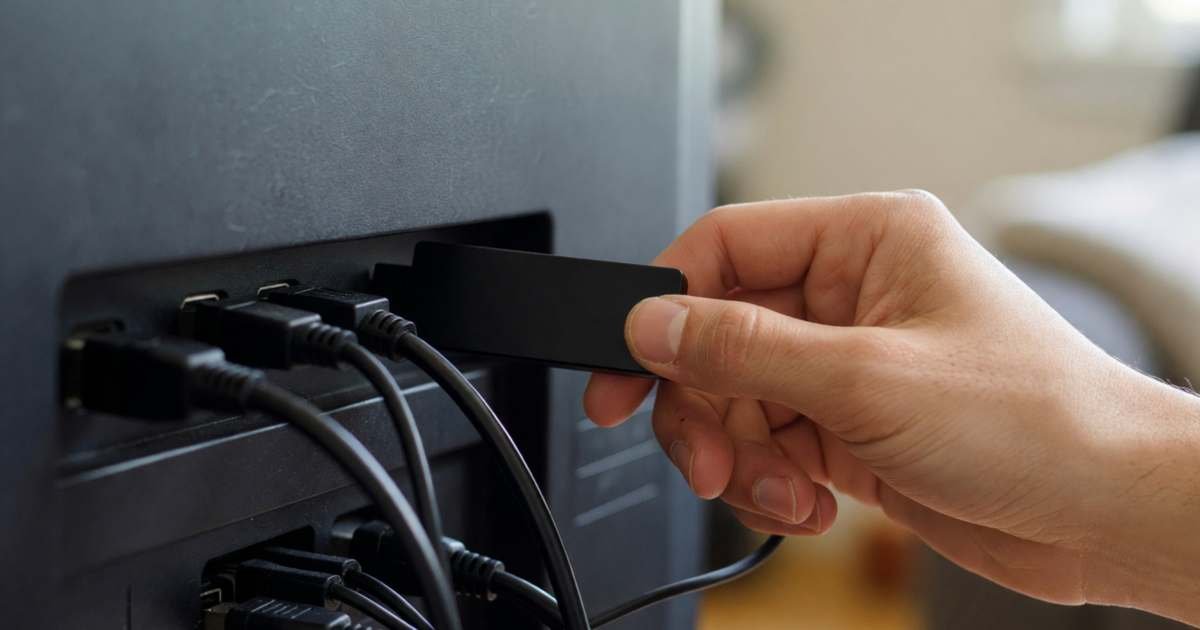 Close-up of a hand plugging a small plain streaming stick into the HDMI port on the back of a flat-screen TV, illustrating streaming sticks vs smart TV apps in Canada.