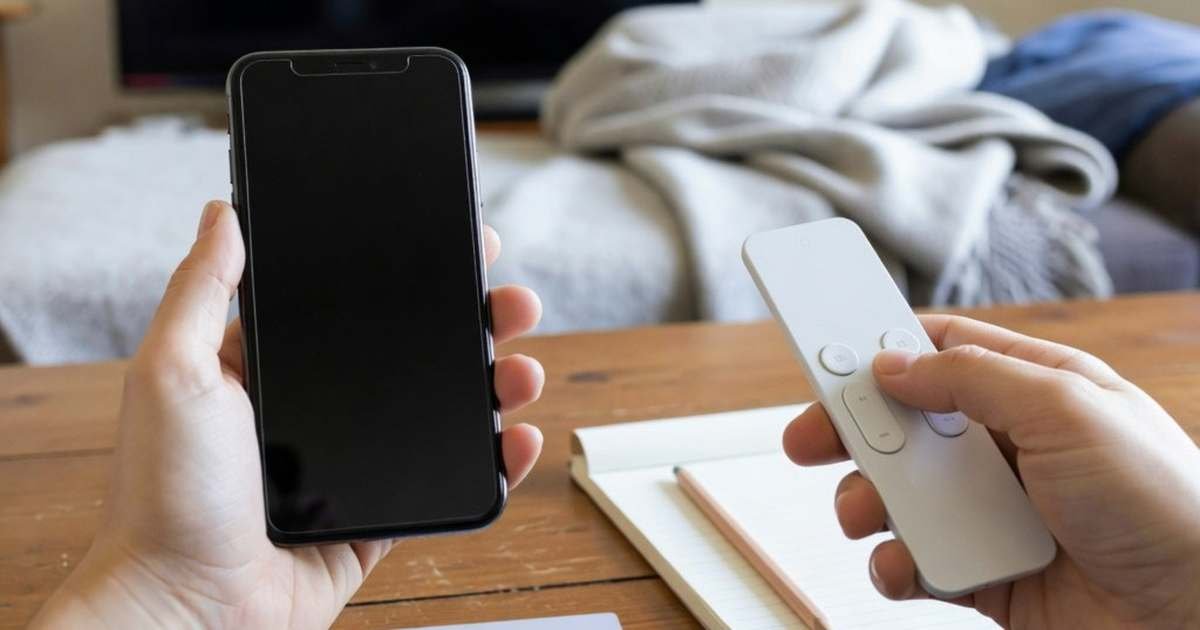 Hands holding a phone and a TV remote beside a blank payment card and notepad in a Canadian living room, illustrating a household subscription audit of streaming services and apps.