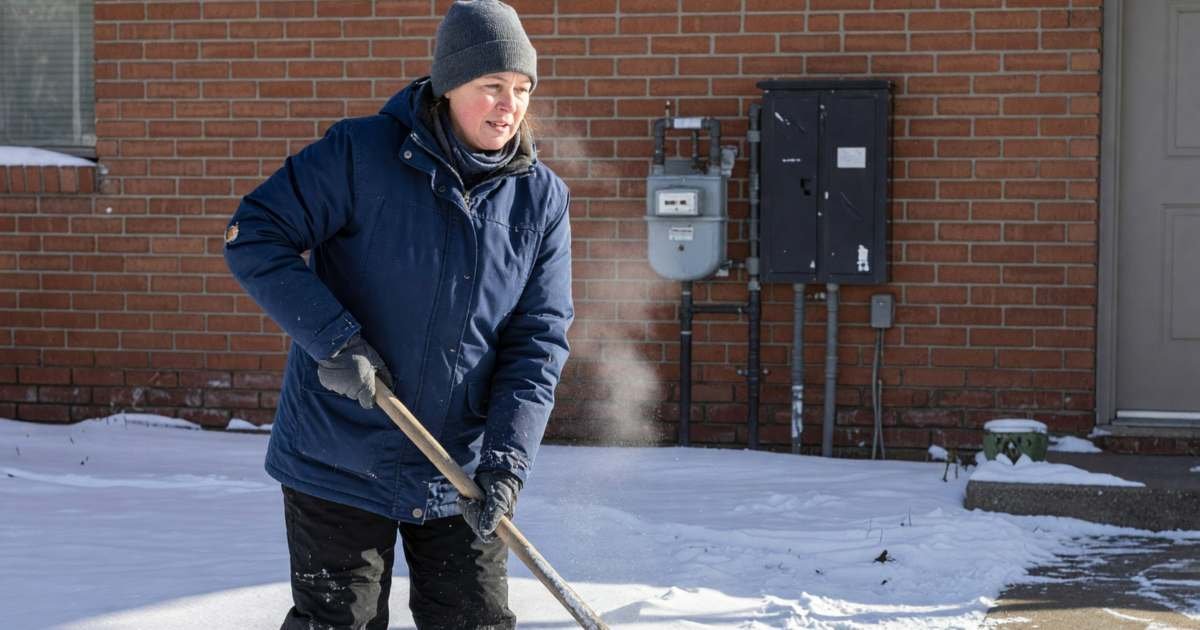 A woman shovelling snow on her front walk with a gas meter and electrical panel on the house wall behind her, illustrating switching electricity or gas retailers in Alberta.