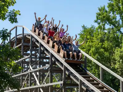 A wooden roller coaster against a summer sky with riders on the first drop.