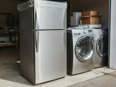 A new stainless refrigerator, washer and dryer still in plastic wrap on a garage floor.