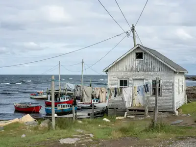 A wooden coastal house with clothesline and power lines leading to it beside the ocean.