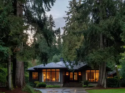 A West Coast house with large windows at dusk, lights on inside, tall cedar trees around it.