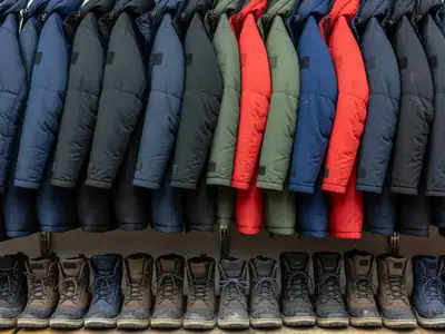 A long rack of heavy winter parkas and a low shelf of winter boots in a quiet clothing store.