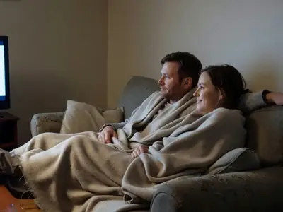 A couple on a sofa under a blanket watching TV at night, seen from behind.
