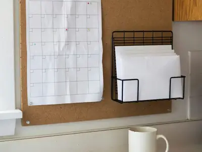 A paper wall calendar with a blank grid pinned on a kitchen bulletin board, some squares marked with coloured dots, beside a few envelopes in a wire organiser.