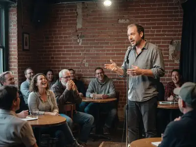 A comedian on a small club stage under a spotlight, audience laughing at small tables.