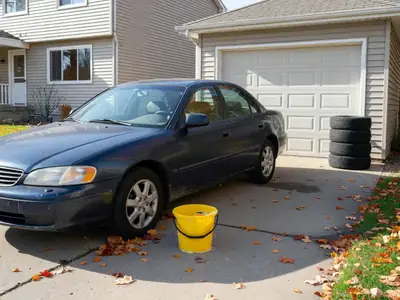 A mid-size family car parked in a driveway beside a house, with a car wash bucket and a set of seasonal tires stacked by the garage.