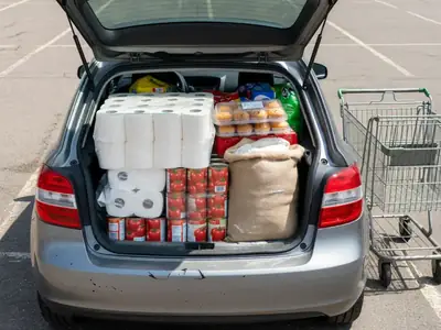 A car hatchback open in a parking lot, packed with bulk groceries: a huge pack of toilet paper, cases of canned tomatoes, a sack of flour and a tray of muffins.