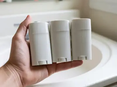 A hand holding three plain deodorant sticks side by side over a bathroom counter.