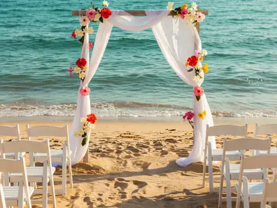 A small beach wedding arch draped with white fabric and flowers facing the ocean, rows of white chairs.