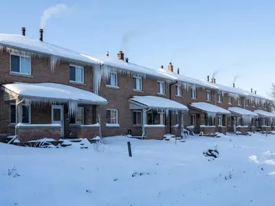 A row of identical townhouses under heavy snow with smoke from vents and long icicles.