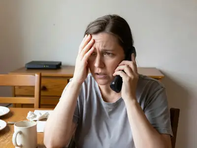 A frustrated woman on the phone at her dining table, rubbing her forehead.