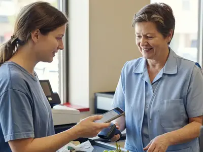 A woman at a checkout showing her phone to a friendly cashier who is scanning a bunch of grapes.