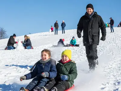 Two laughing children riding a red plastic sled down a snowy park hill while their dad jogs alongside, other families scattered on the slope.