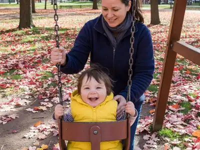 A nanny pushing a toddler on a playground swing.