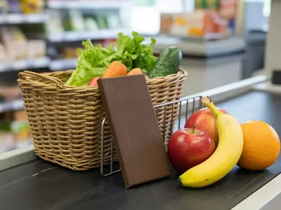 A chocolate bar sitting back on a rack beside fruit and a basket of vegetables at a grocery checkout.