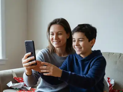 A mother handing her twelve-year-old son a smartphone with a dark screen, the boy grinning.
