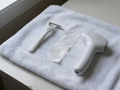 A razor, a wax strip and a small laser hair removal handset laid out on a white towel.