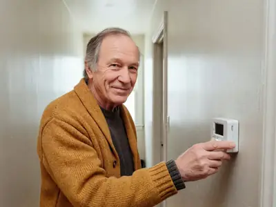 An older man in a warm cardigan turning down a plain wall thermostat in his hallway.