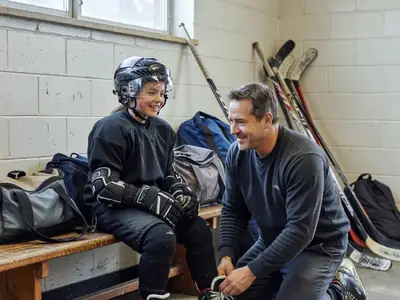 A young girl in full hockey gear sitting on a dressing-room bench while her dad tightens her skates.