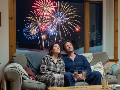 A couple in pajamas watching fireworks through a window at midnight with sparkling drinks.