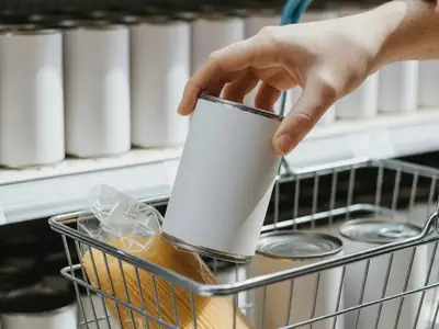 Close-up of a hand placing a plain unlabelled can of tomatoes into a shopping basket already holding pasta and beans.