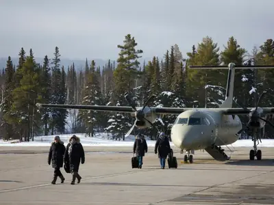 A small regional airport with a turboprop plane on the tarmac and boreal forest beyond.