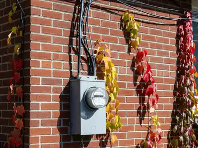 An electricity meter on the brick side wall of a house with wires leading up to the roofline.