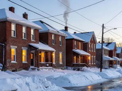 A row of older brick houses on a snowy street at dusk, warm lights glowing in every window and smoke rising from a chimney.