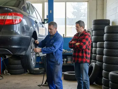 A mechanic in a tire shop mounting a winter tire on a car lifted on a hoist while the owner looks on.
